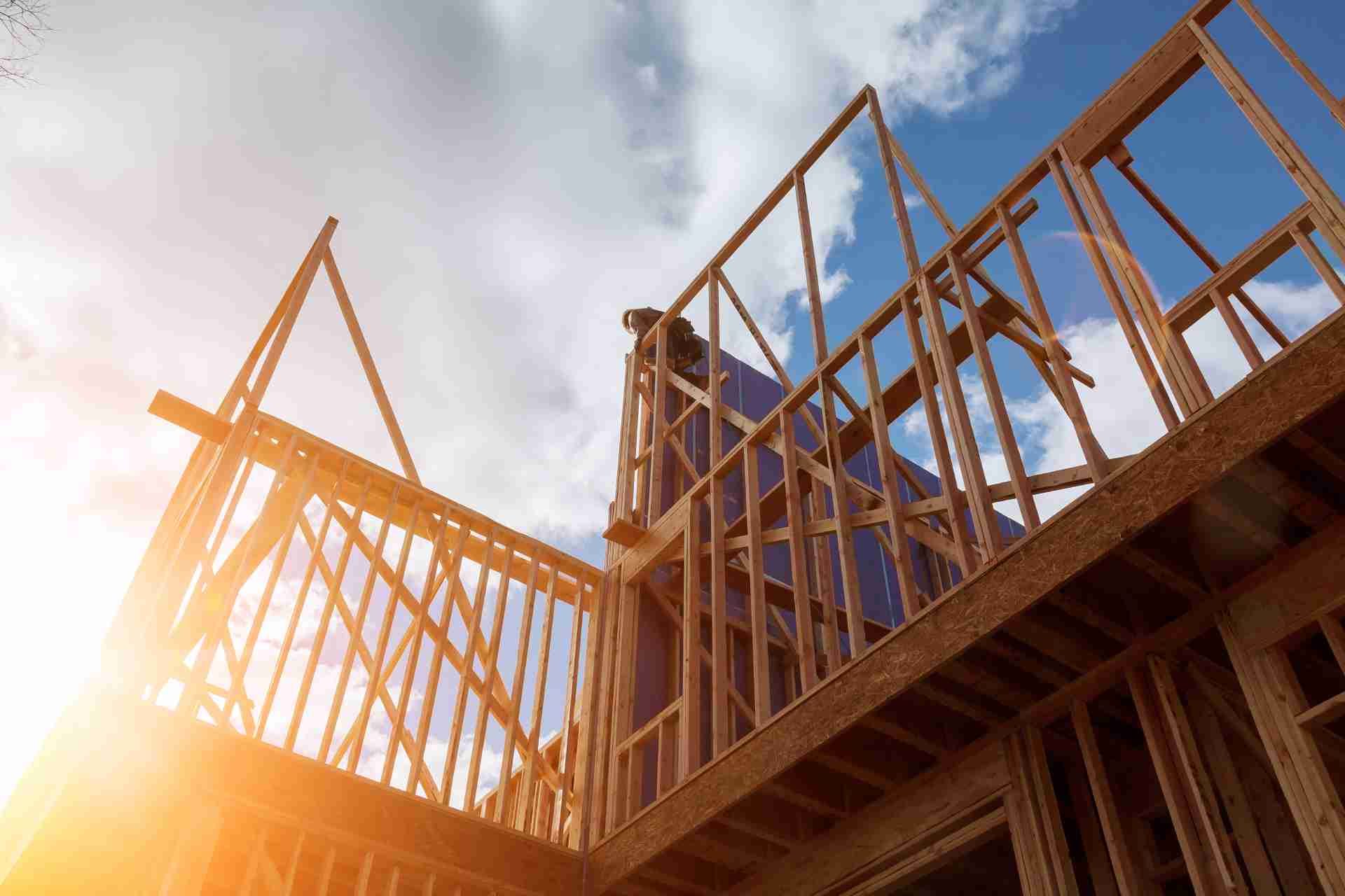 A wooden house frame under construction with a worker on top and the sun shining through the structure