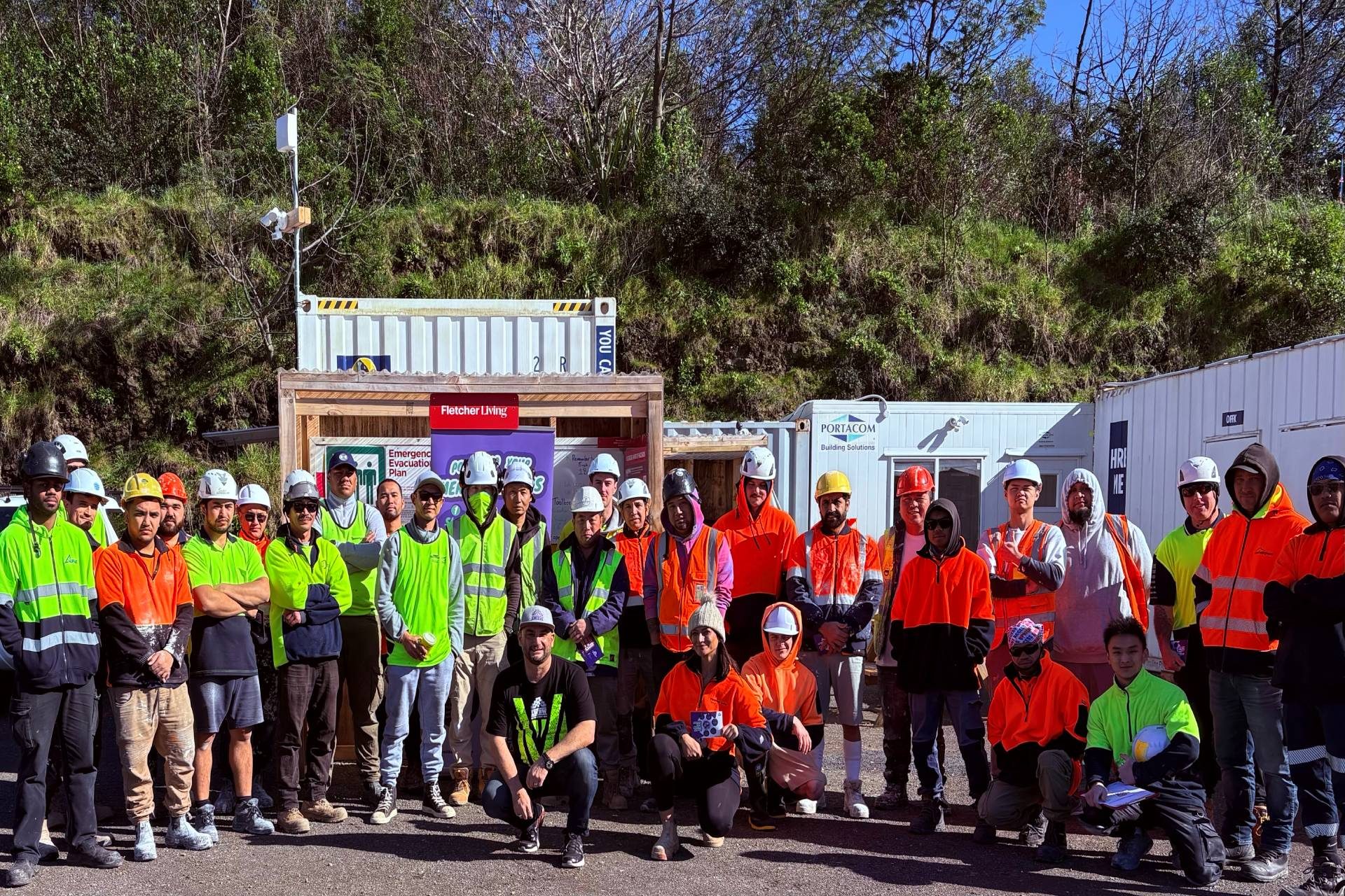 A large group of construction workers wearing safety vests and helmets pose together outdoors at a job site.